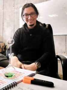 El profesor Nicolás Fernández está sentado detrás de un escritorio, sobre éste hay un cuaderno y un matraz.